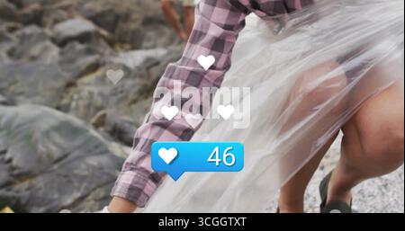 Man wearing flannel wrapping film around her on pebbly beach, with white hearts, blue badge 46 Stock Photo