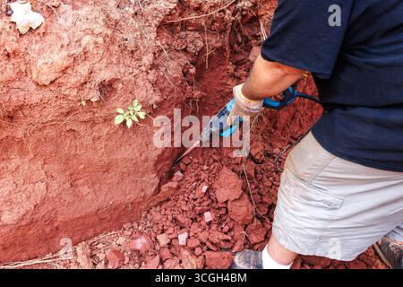 Work man uses power jackhammer tool to dig into red soil in construction outdoor area Stock Photo