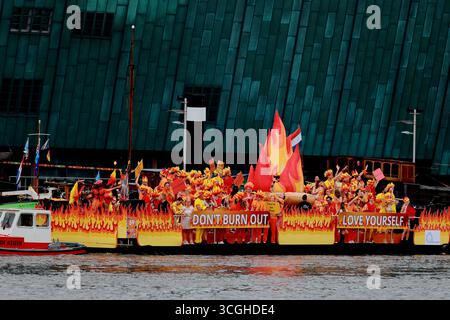 Orange flame themed boat reading 'Don't Burn Out, Love Yourself' carries performers along the Pride Amsterdam Canal Parade in Amsterdam, Netherlands. Stock Photo