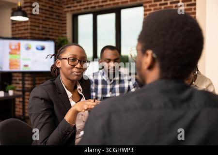 Female manager talks through marketing strategies with black male employee, asking questions and analyzing financial report. Businesswoman discussing project updates with colleague in modern office. Stock Photo
