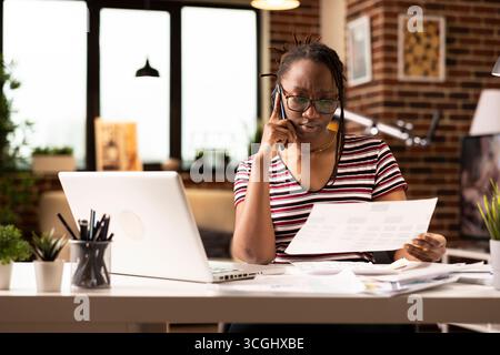 African american freelancer reviewing important papers during serious phone call with business client. Focused black woman with documents in hand, talking with coworker through mobile device. Stock Photo