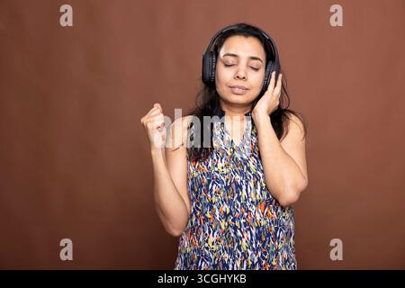 Beautiful woman wearing wireless headphones, deeply immersed in music. Female model listens with closed eyes and raised fist, enjoying the rhythm and beat against isolated background. Stock Photo