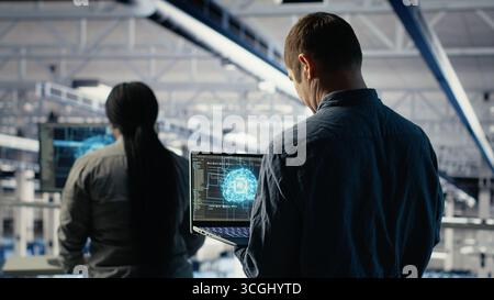 Data center system administrators investigate neural networks configuration, adjusting for optimal function. Server hub employees use laptop to check deep learning error reports, camera B Stock Photo