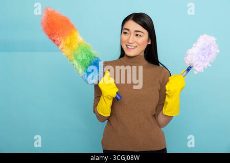 Happy asian woman wearing yellow rubber gloves holds rainbow and white duster, smiling confidently while posing for portrait against blue background. Housekeeping and hygiene concept. Stock Photo