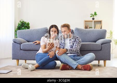 Happy positive husband and wife sharing phone screen at home together, sitting on warm floor Stock Photo