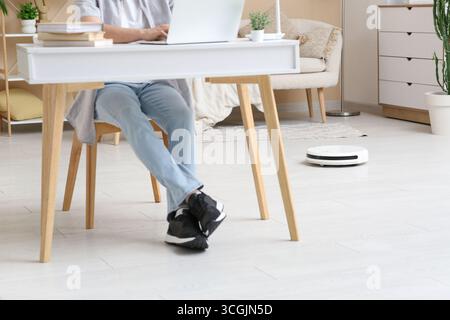 Man using laptop at table with modern robot vacuum cleaner on floor in home office Stock Photo