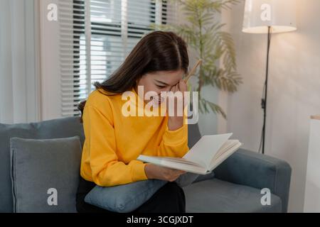 Young Asian woman in yellow sweater sitting on sofa and smiling while writing notes in notebook. Relaxed atmosphere in cozy living room. Concept of le Stock Photo