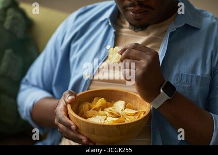 Young adult Black man sitting indoors holding wooden bowl eating potato chips with right hand wearing smartwatch on left wrist, focusing on snack in relaxed setting Stock Photo