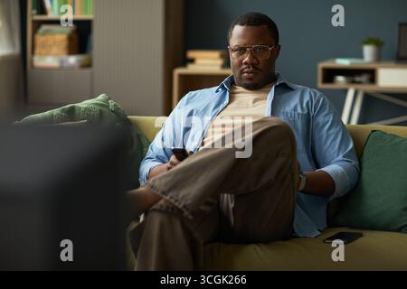 Portrait of Black man sitting on sofa holding remote control and watching television, wearing glasses and casual clothing, smartphone resting beside him on cushion Stock Photo