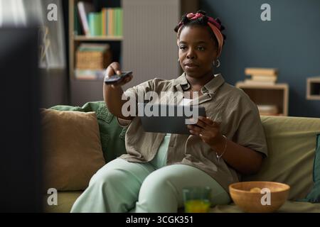 Young adult Black woman sitting on sofa holding digital tablet in one hand and using remote control with other hand, watching television with focused expression Stock Photo