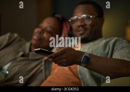 Black man holding remote control sitting next to Black woman relaxing on couch watching television together, both appearing comfortable and engaged in activity Stock Photo
