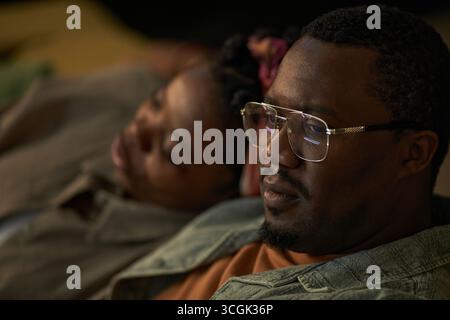 Black man with glasses sitting closely with Black woman resting head on his shoulder, both appearing relaxed, man looking away from camera, indoor moment captured Stock Photo