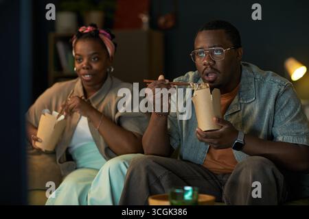 Black man and Black woman sitting on couch eating takeout food with chopsticks, watching television together, both appearing as young adults, focused on screen Stock Photo
