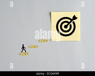 Businessman climbs paper clips toward target drawn on sticky note, representing business goal setting and achieving objectives. Stock Photo