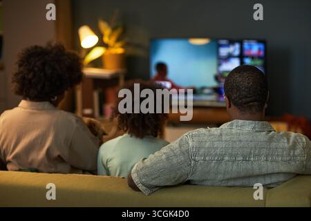 Black family sitting on sofa watching television together, showing back view of woman, man, and child with curly hair focusing on screen in living room Stock Photo