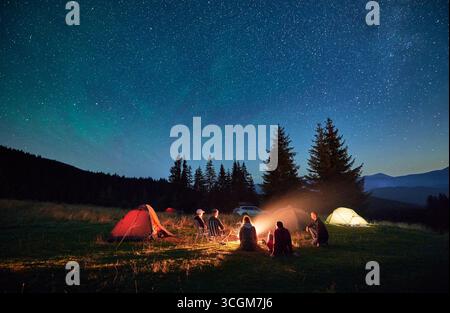 Group of friends hikers having a rest around campfire, surrounded by glowing tents under star-filled sky. Serene mountain backdrop and night sky create magical and peaceful atmosphere in wilderness. Stock Photo
