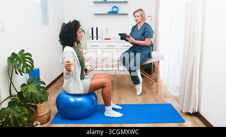 A Senior Physiotherapist Guides a Young Black Client Through Strength Exercises on an Exercise Ball, Focusing on Recovery Stock Photo