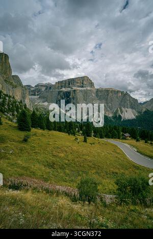 Sella Group in the Dolomites with dramatic limestone cliffs rising above alpine meadow and winding road under cloudy mountain sky Stock Photo