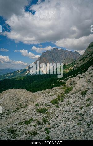 Dolomites mountain peak above forested valley with rocky limestone slope in the foreground under partly cloudy blue summer sky Stock Photo