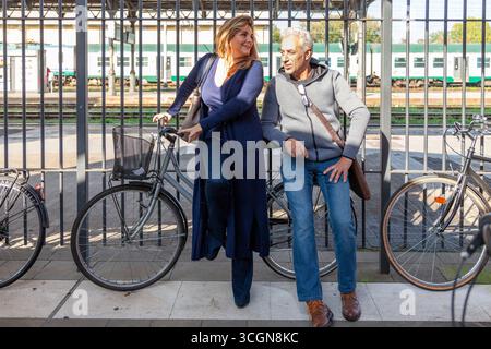 A mature woman and man talk while leaning on their bicycles in the train station parking lot in front of a train and a metal fence. Stock Photo