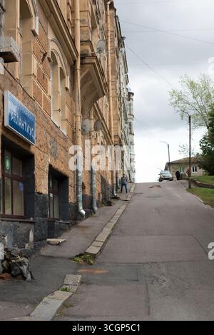 viborg, saint petersburg, russia, 26.08.2019 An old man walking down a steep, worn asphalt road with a car parked at the top of the hill, leading to u Stock Photo