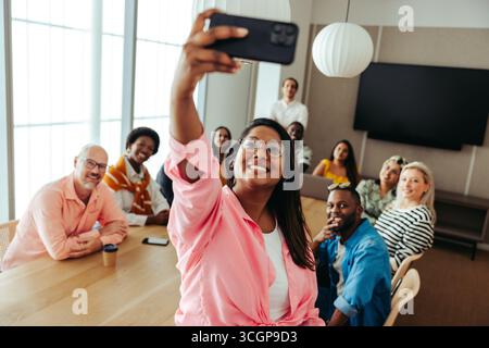 A diverse group of colleagues gathers around a table in a modern office to capture a joyful selfie. Everyone smiles, showcasing teamwork and camarader Stock Photo