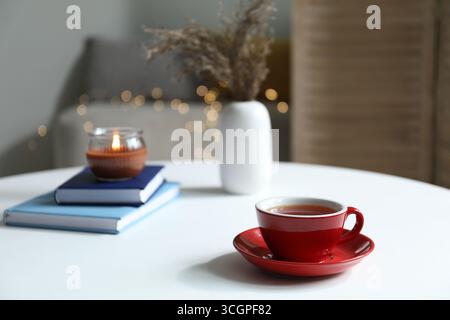 Cup of tea, books and burning candle on white table against blurred lights Stock Photo