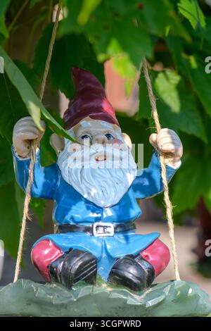 A colorful garden gnome with a red hat and white beard swings from a rope under bright green leaves. Stock Photo