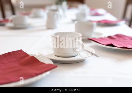 Empty coffee cups and plates with red napkins on a large table. Objects related to tea time and eating cake with the family. Decoration at an event. Stock Photo