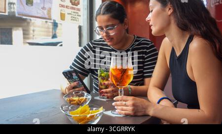 Two Young Women Enjoying Refreshing Aperitif Drinks and Snacks Together While Checking Their Phones in a Bright Outdoor Cafe Setting on a Sunny Day Stock Photo