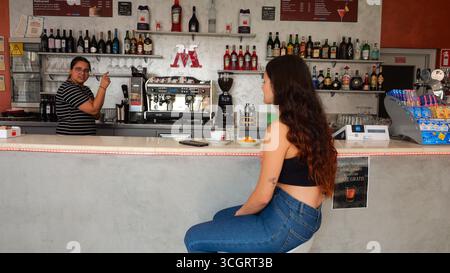 Young woman wearing blue jeans and black top sits at a bar counter receiving a drink from a barista wearing a black and white polka dot shirt in a coz Stock Photo