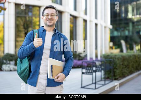 Confident young man holding a notebook and backpack standing outdoors. Advocating success and positivity with a cheerful gesture in an urban contemporary environment. Stock Photo
