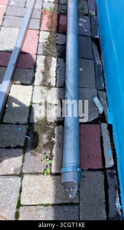 A long metal pipe rests on a paved surface, surrounded by small plants and dirt. The pavement is colorful, showing various blocks and a blue wall near Stock Photo