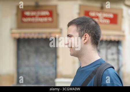 A man with a blue shirt and black backpack is smiling in front of a building with a red sign Stock Photo