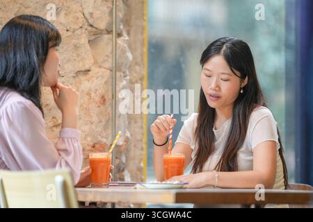 Two women sitting at a table with orange juice and a straw in their drinks. One of the women is looking at the other Stock Photo