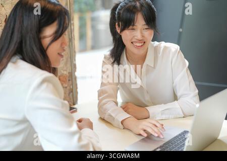 Two women are sitting at a table, one of them is typing on a laptop. They seem to be having a conversation or working together on a project. Scene is Stock Photo