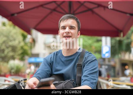 A man is sitting under a red umbrella with a blue shirt on. He is holding a cell phone in his hand Stock Photo