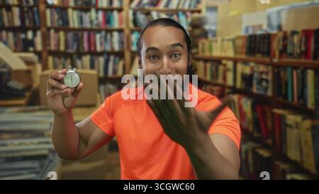 Man holds stopwatch and reaches hand forward in library building while smiling and showing stopwatch to camera; urgency time pressure. Stock Photo