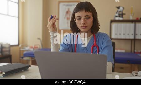 Woman holding pen at laptop in clinic with red stethoscope hanging on neck and anatomical chart behind; concentration. Stock Photo