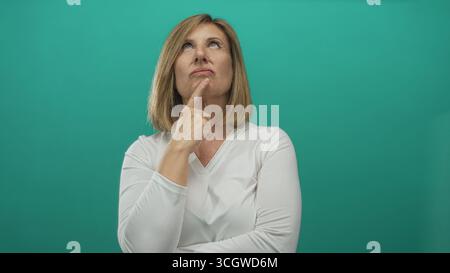 Middle-aged woman wearing white long sleeve shirt resting chin on finger and gazing upward against teal studio wall; thinking. Stock Photo