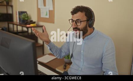 Hispanic man with beard wearing headset in office setting, engaging through computer for virtual communication, emphasizing modern workspace atmospher Stock Photo