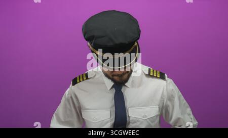 Young hispanic man in pilot uniform with cap looking down against pink background Stock Photo