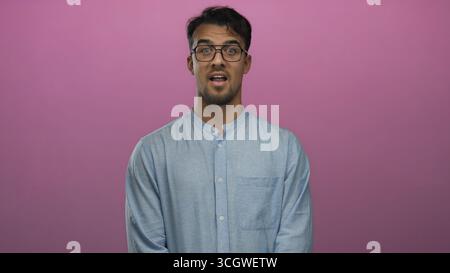 Young hispanic man wearing glasses and blue shirt, expresses surprise and excitement over pink background using facial expressions and gestures. Stock Photo