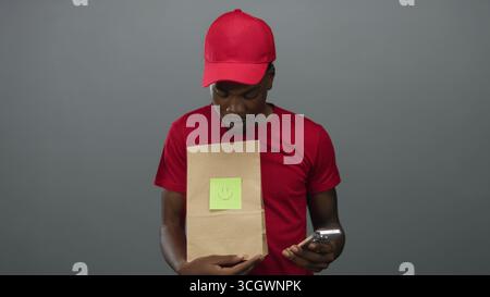 Young man holding smartphone and paper bag with smiley note while looking down in gray studio; curiosity. Stock Photo