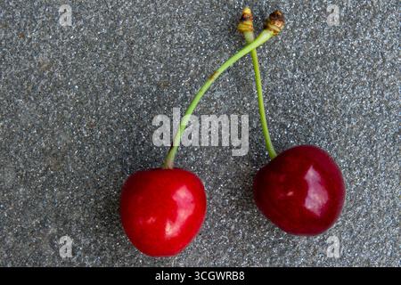 Two vibrant red cherries with stems positioned closely together on a textured gray stone surface, illuminated by soft lighting. Stock Photo