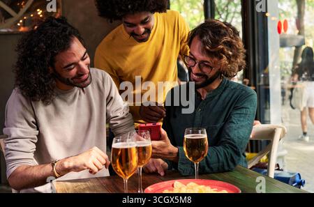 Group of man friends in a bar having fun, drinking beer, using one phone to watch social media. Stock Photo