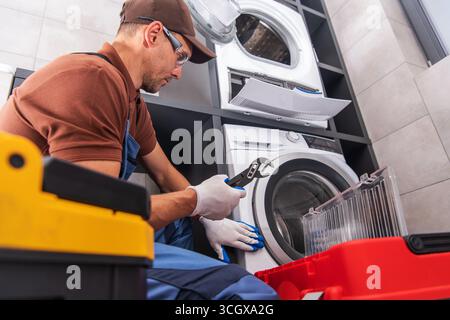 A technician is working on repairing a washing machine in a stylish laundry room, focused on fixing the unit with various tools near him. Stock Photo