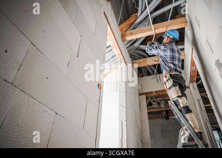 A construction worker is using a ladder to install electrical wiring in the ceiling of a new building. The workspace features exposed beams and materi Stock Photo