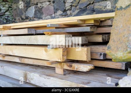 A neat stack of new and old wooden planks rests against a weathered stone wall, highlighting construction materials and textures. Stock Photo