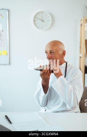 Senior elderly male doctor recording audio message to a patient using smartphone mic. copy space Stock Photo